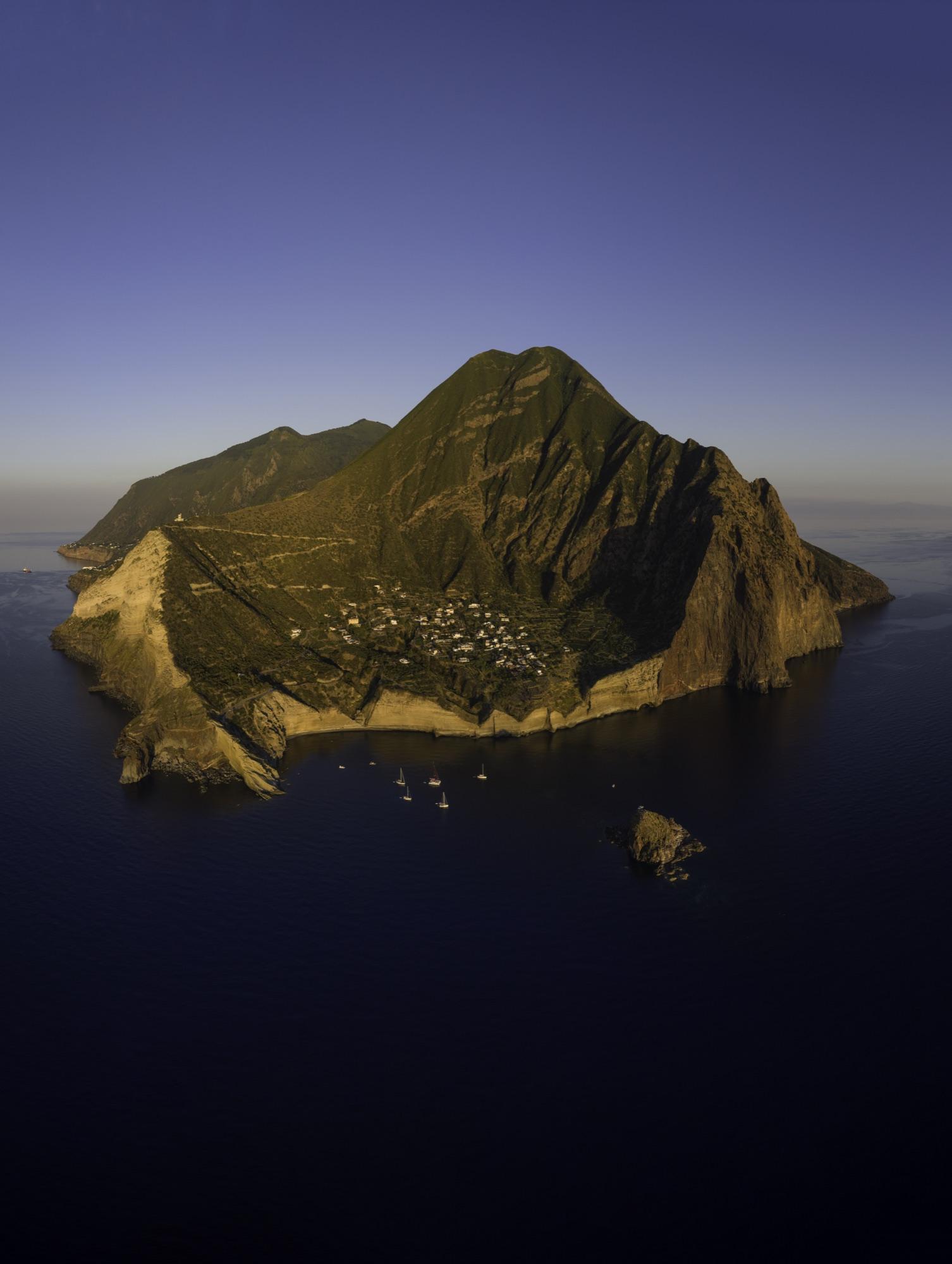 Aerial view of a volcanic Aeolian island at golden hour with sailboats moored offshore