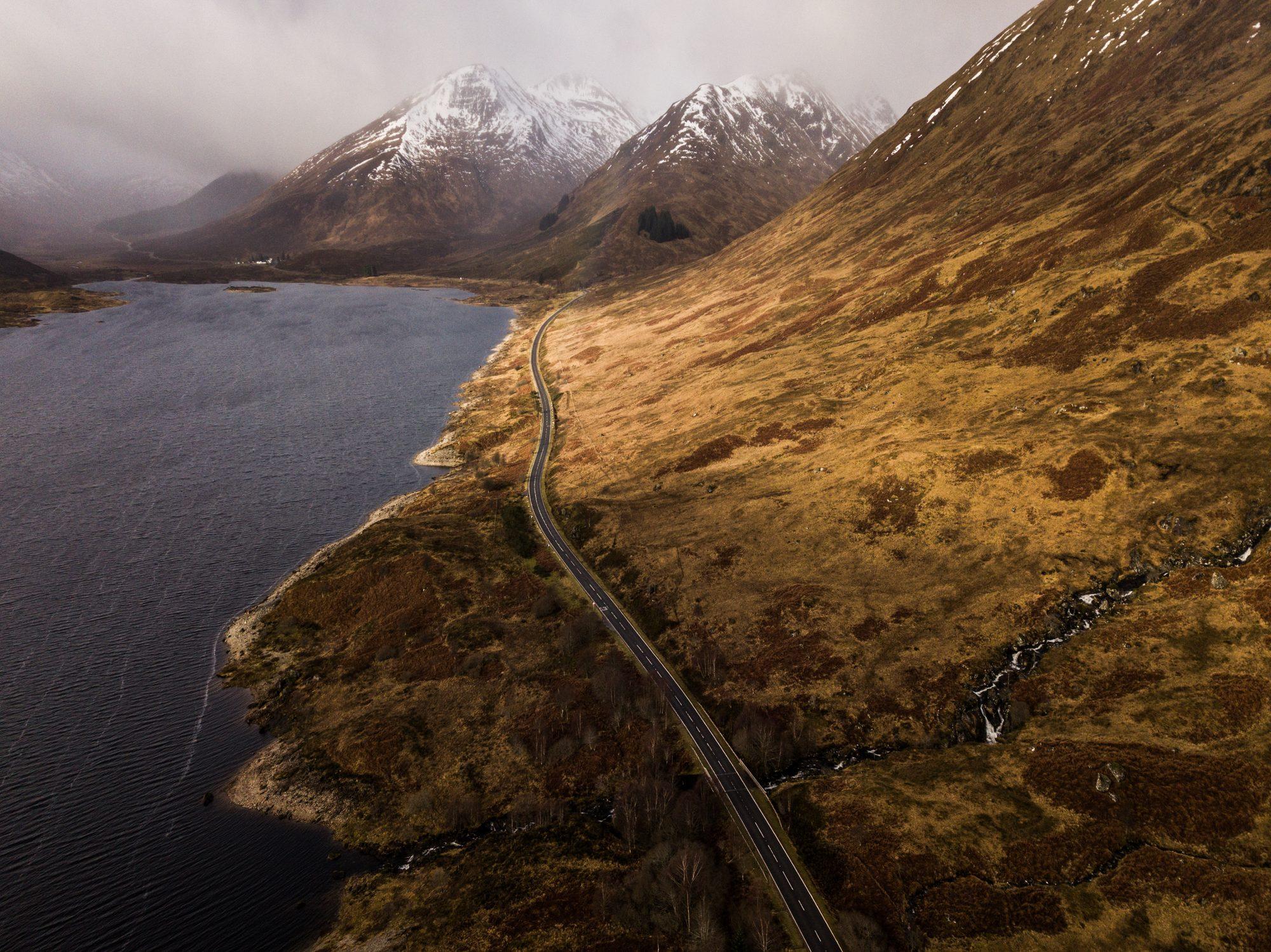 Aerial view of a winding road along a loch beneath snow-capped Highland mountains