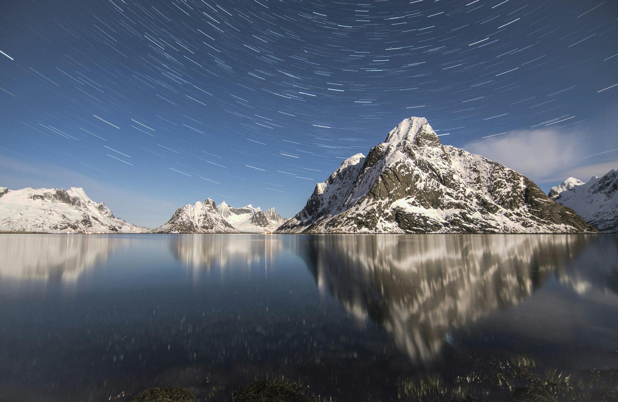 Star trails circling above a snow-capped peak and lake